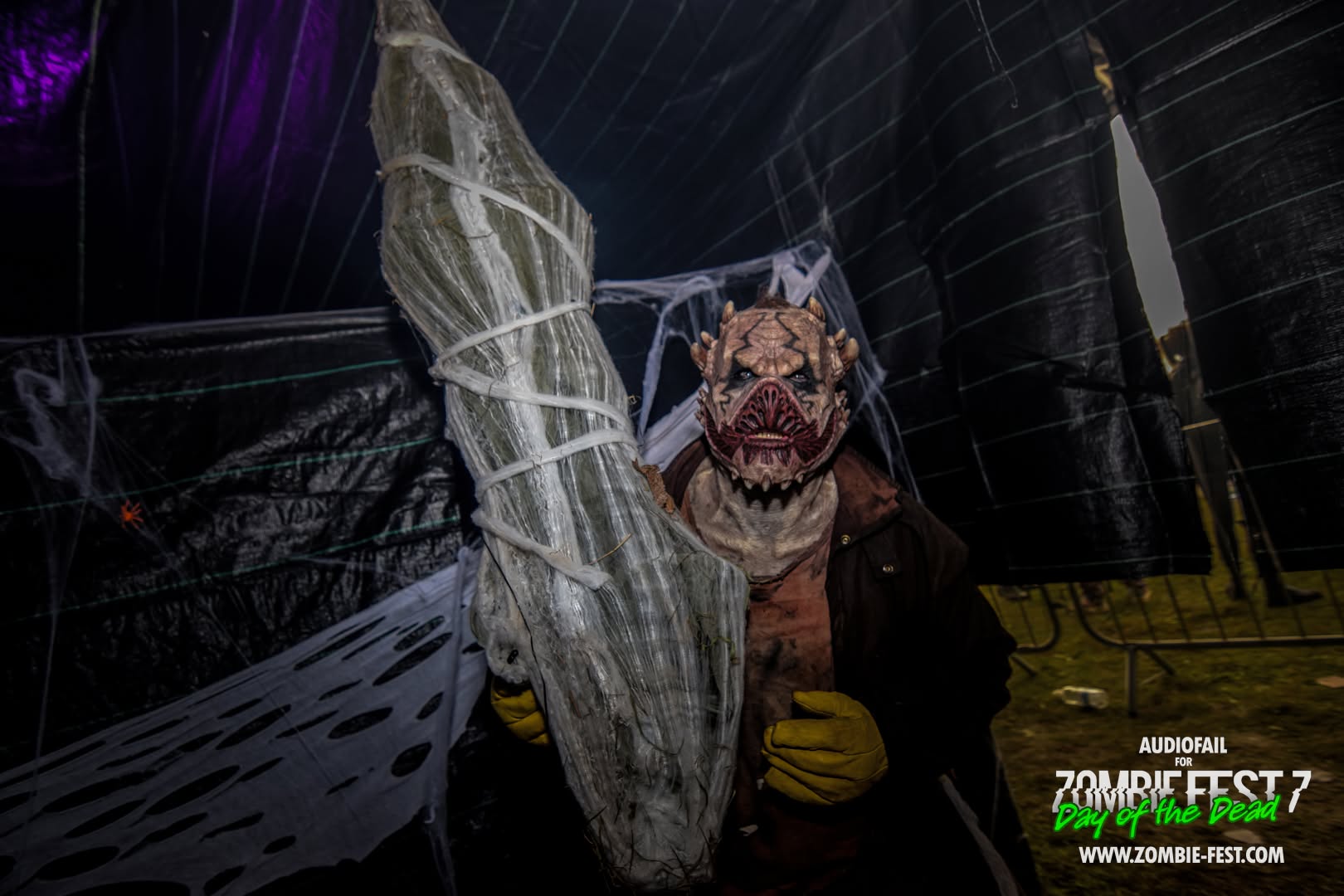 Costumed creature with fanged mask carrying a wrapped cocoon body inside a cobwebbed tent at Zombie Fest 7