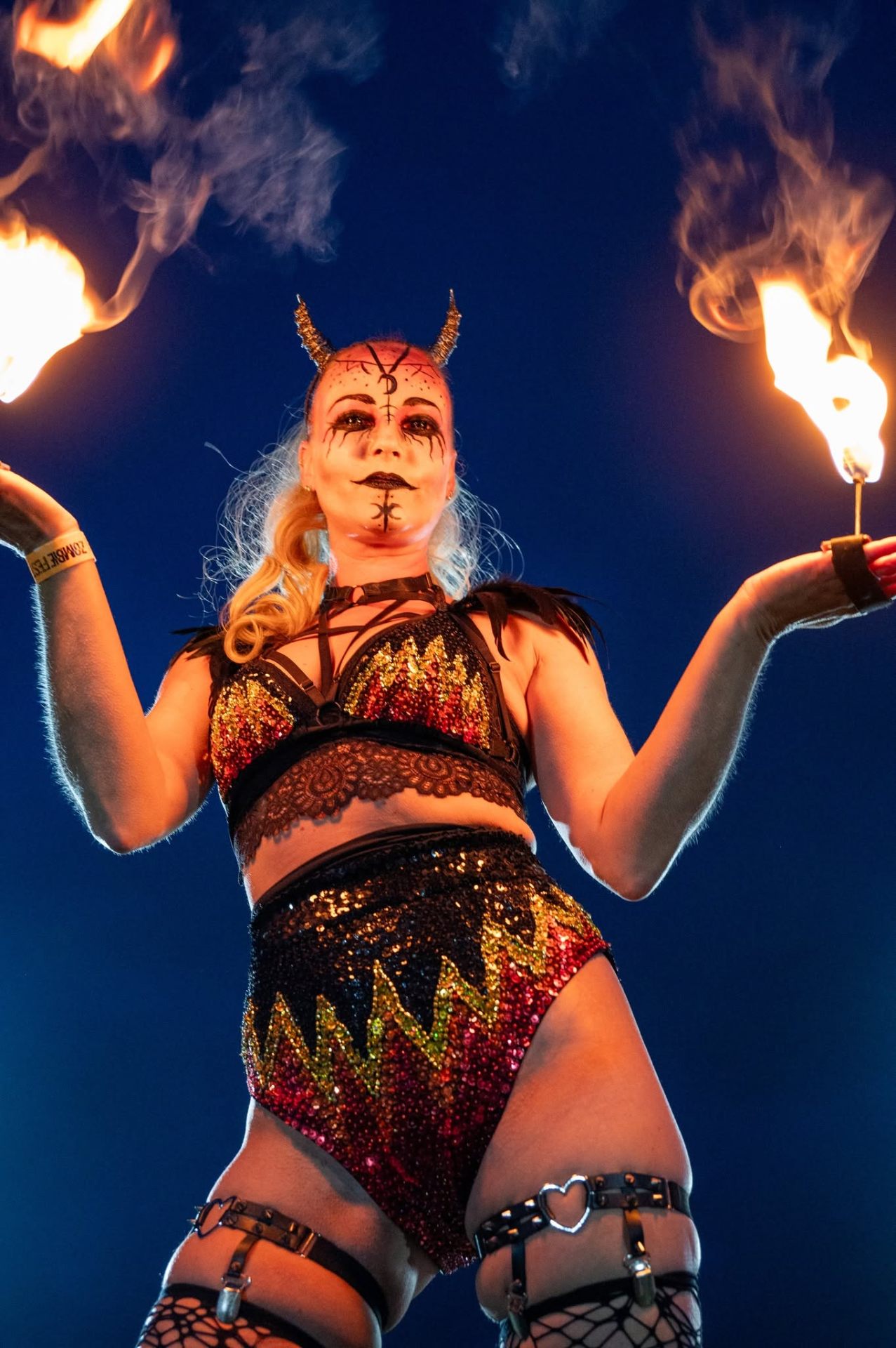 Fire performer with horned demon makeup holding two lit torches against a night sky at Zombie Fest