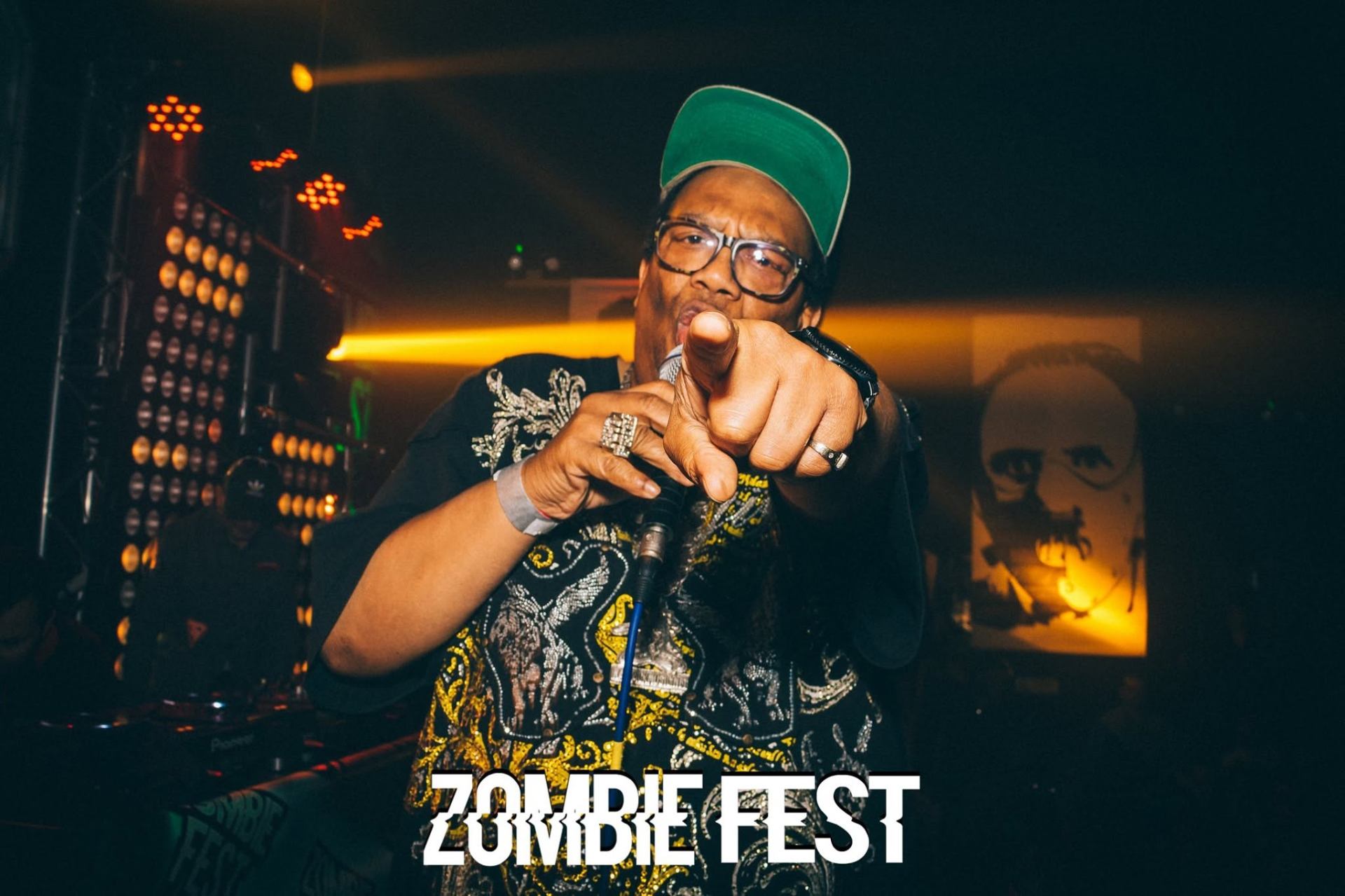 Ratpack MC pointing at the camera on stage under stage lights at Zombie Fest